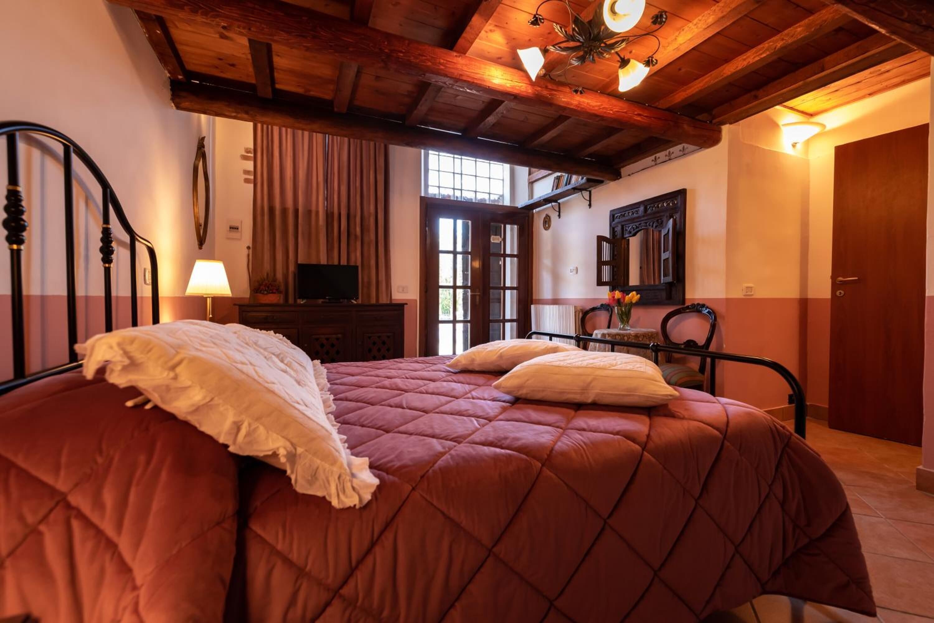 A bedroom with a large bed, wooden ceiling beams, a television, and a window.