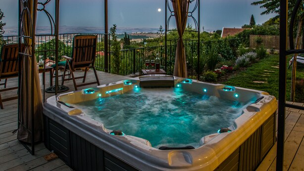Un jacuzzi en una terraza de madera con sillas y una mesa, con vista a un jardín y un paisaje lejano.