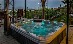 Un bain à remous sur une terrasse en bois, agrémentée de chaises et d’une table, offrant une vue sur un jardin et un paysage lointain.