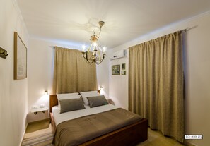 A neatly arranged bedroom with a bed, bedside lamps, a chandelier, and curtains. - Naomi's (Rosh Pinna)