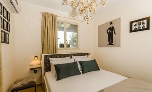 A bedroom with a bed, a window with curtains, a wall-mounted picture, a wall-mounted lamp, and a small table with a chair.