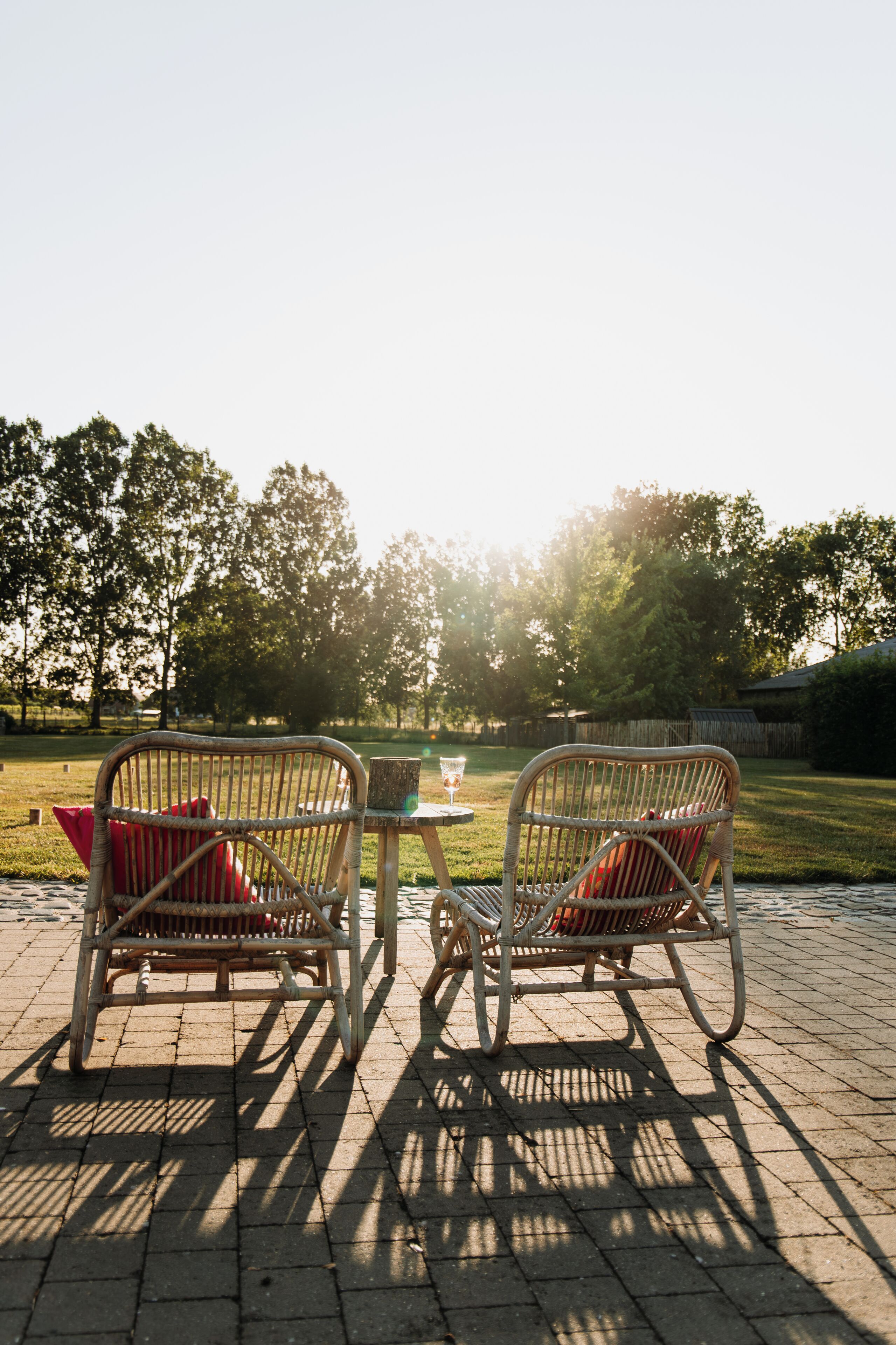Two wicker chairs with red cushions and a small round table with a glass on it, set on a paved patio with a view of a grassy area and trees in the background.