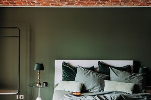 Un dormitorio con pared de ladrillo, cama con muchos cojines, mesita de noche con lámpara y estante fijado a la pared.