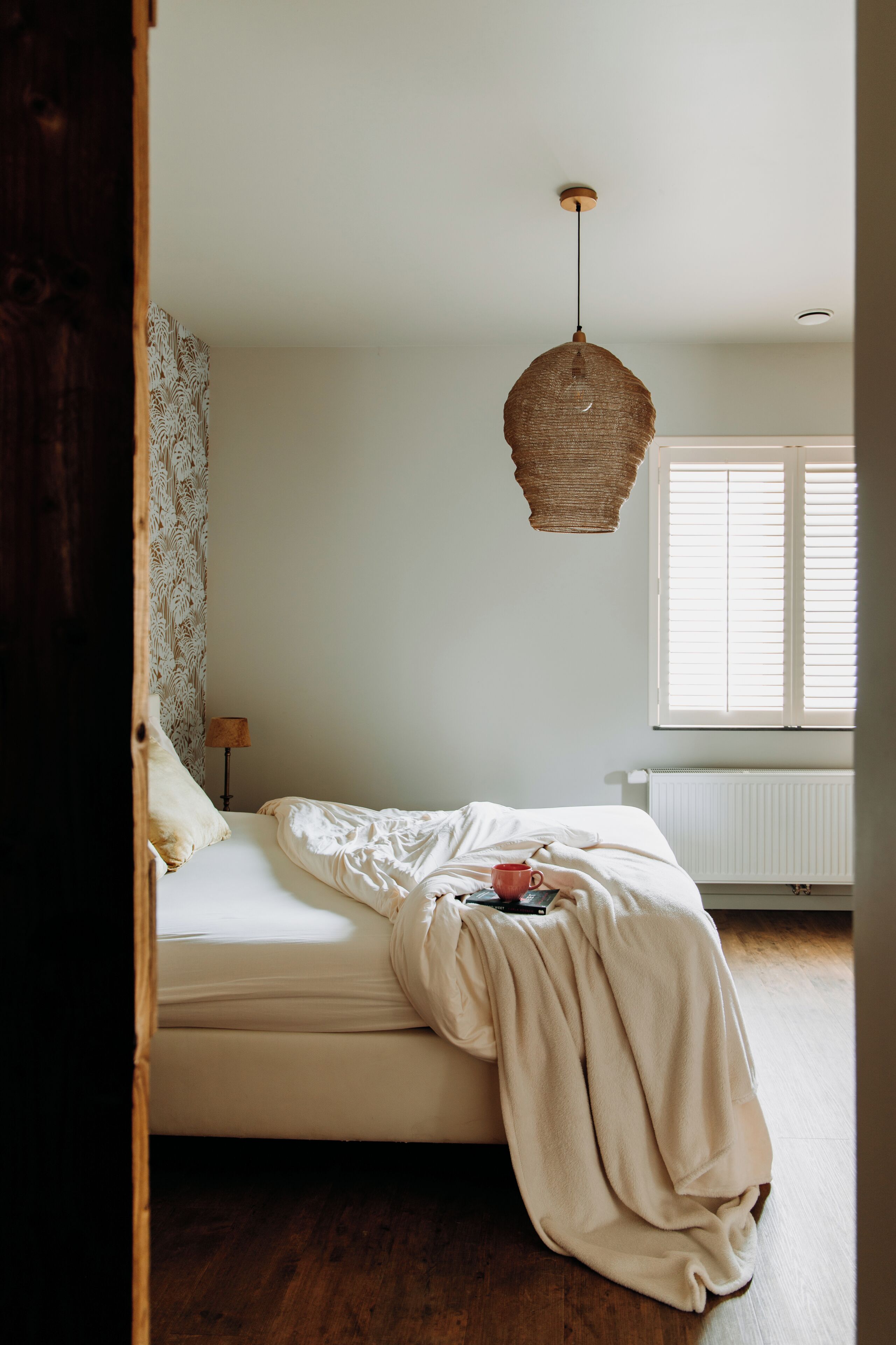 Um quarto com uma cama, luminária pendente, janela com persianas e radiador.
