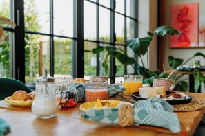 A breakfast table with various food items and drinks, set in a room with large windows and indoor plants. - B&B Casa Cava (Dendermonde)