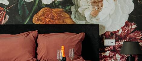 Un dormitorio con un amplio mural floral en la pared, una cama con ropa de cama rosa y una botella de champán sobre la mesita de noche.