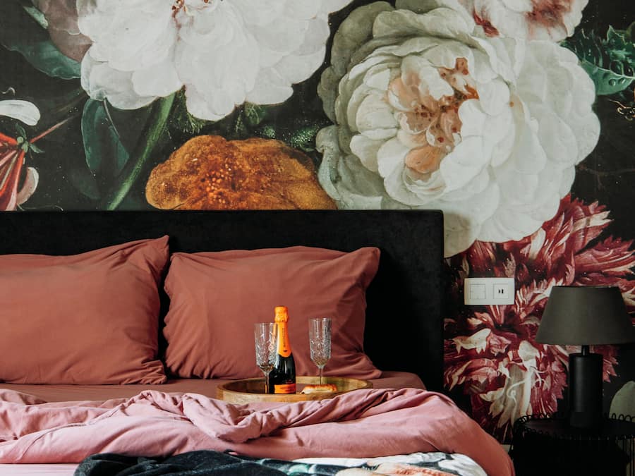 Une chambre avec un grand décor mural floral sur le mur, un lit avec une literie rose et une bouteille de champagne sur la table de chevet.