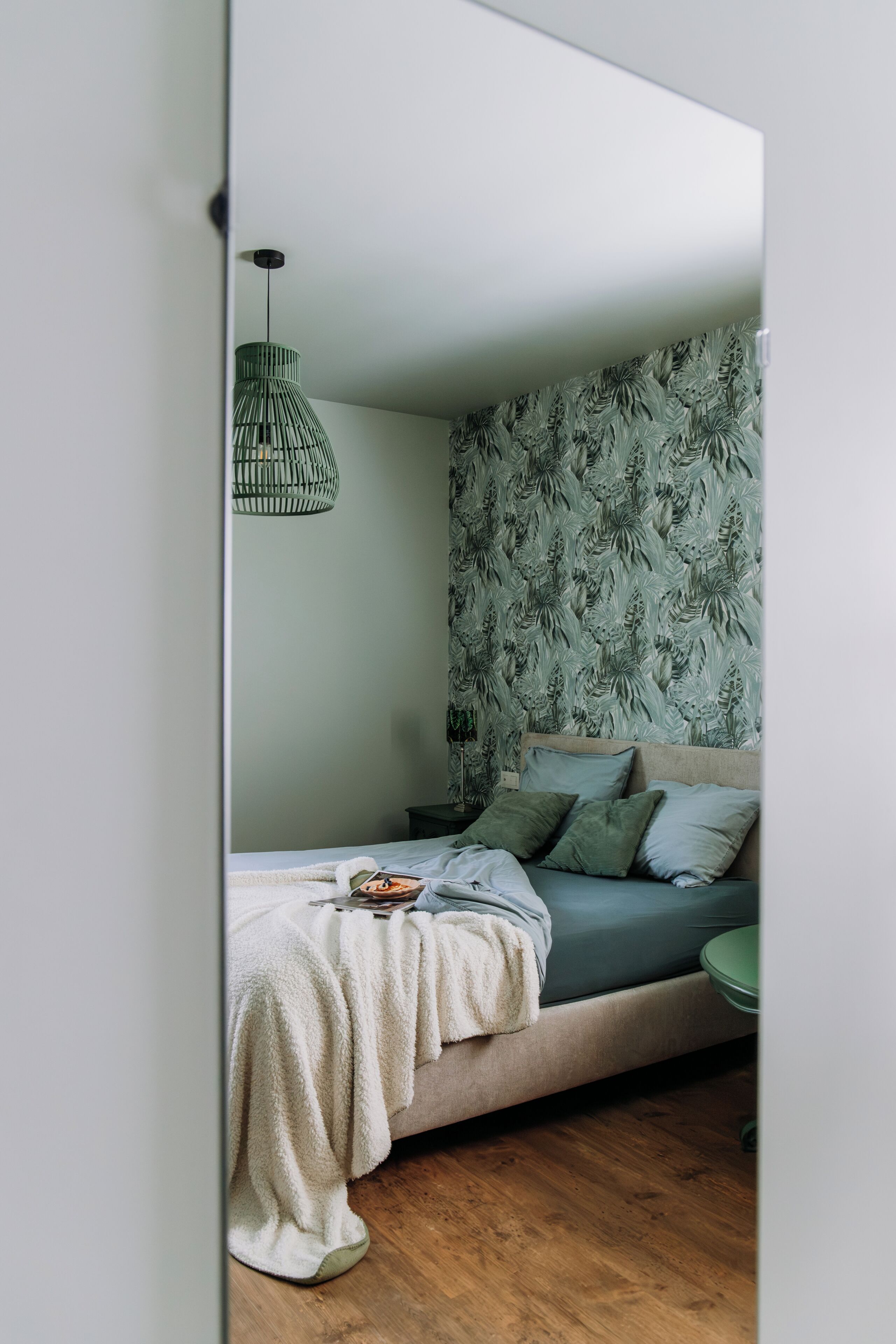 Quarto com cama, almofadas, cobertor, luminária pendente e parede com padrão de folhas.