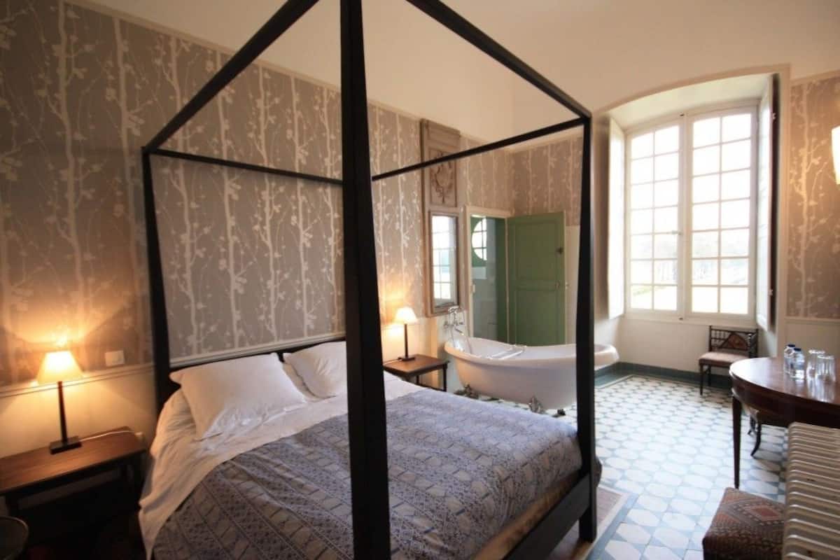 Een bed met vier poster, een bed met een dessin, een vrijstaand bad, een houten tafeltje en een stoel in een kamer met behang met patroon en een betegelde vloer.
