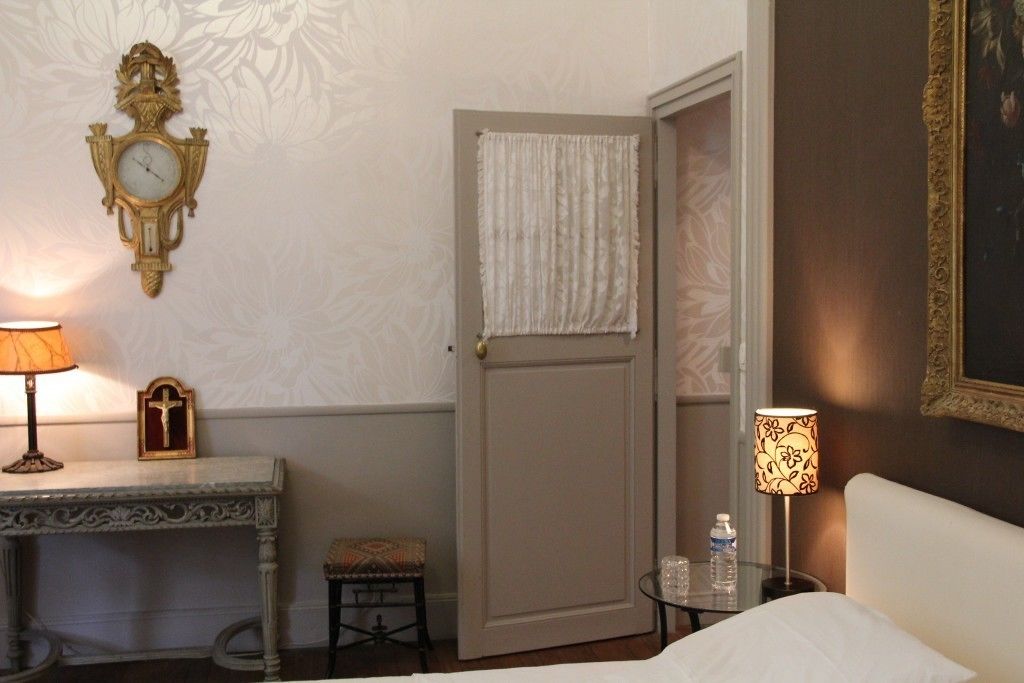 A hotel room with a bed, a nightstand with a lamp, a small table with a cross, a wall clock, and a door with a curtain.