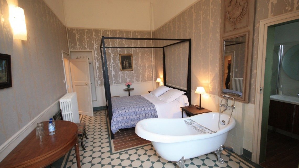 Un dormitorio con cama con dosel, una bañera blanca, una mesita de noche pequeña con lámpara y una puerta que da a otra habitación.