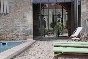 A stone building with a pool, a glass door, and two lounge chairs.