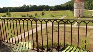 Un balcon doté d’une balustrade en métal, donnant sur un champ verdoyant et sur une petite structure ressemblant à une tour, visible au loin.