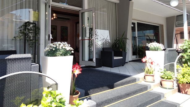 Entrada de hotel con un área exterior cubierta, escalones que conducen a la entrada y plantas en macetas.