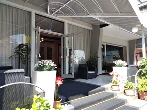 Entrada de hotel con un área exterior cubierta, escalones que conducen a la entrada y plantas en macetas.