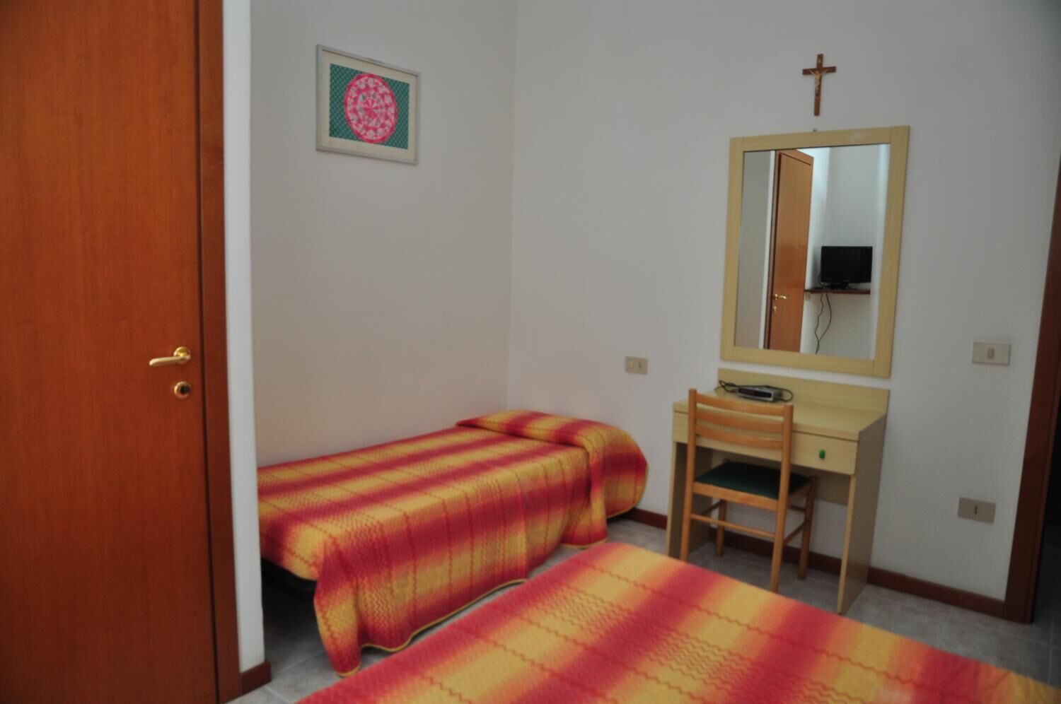 A small, simple room with two single beds, a wooden desk, a chair, a mirror, and a framed picture on the wall.