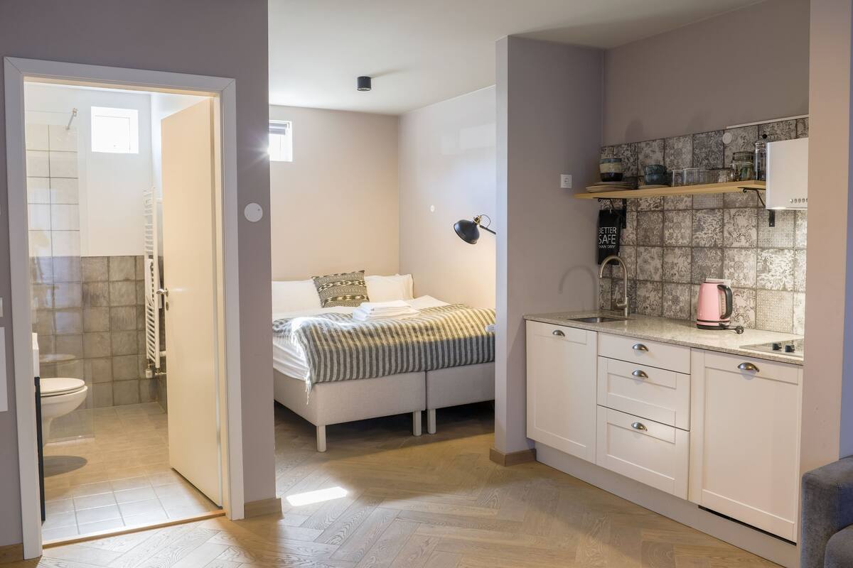 A compact bedroom with a bed, bedside table, and a small window. Adjacent is a kitchenette with a countertop, sink, and a kettle.