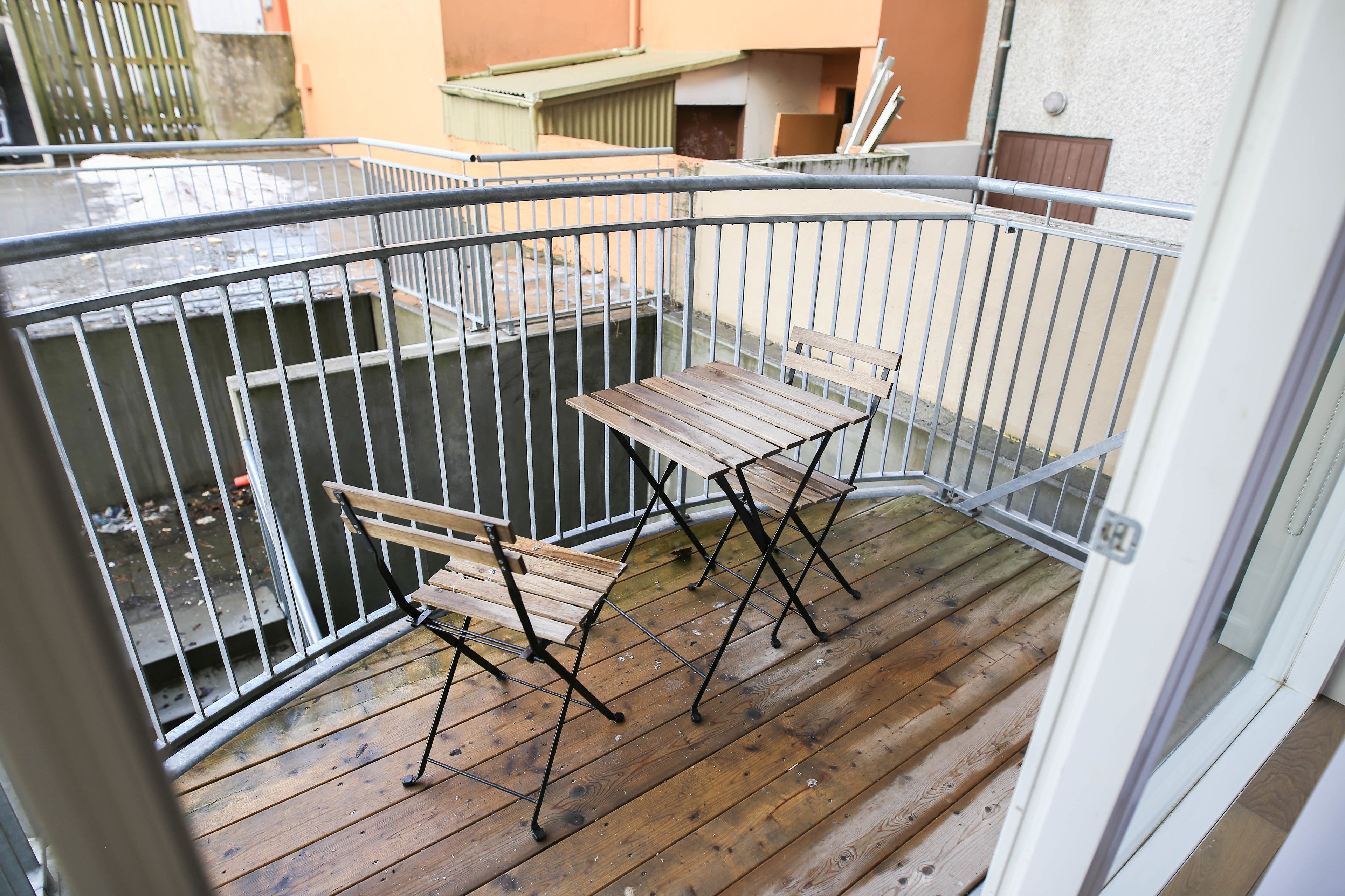 A balcony with a wooden table and chairs, metal railing, and a view of neighboring buildings.