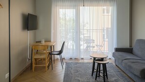 Una sala de estar con un sofá gris, una mesa de comedor de madera con dos sillas, una mesita redonda con un taburete y un televisor montado en la pared.
