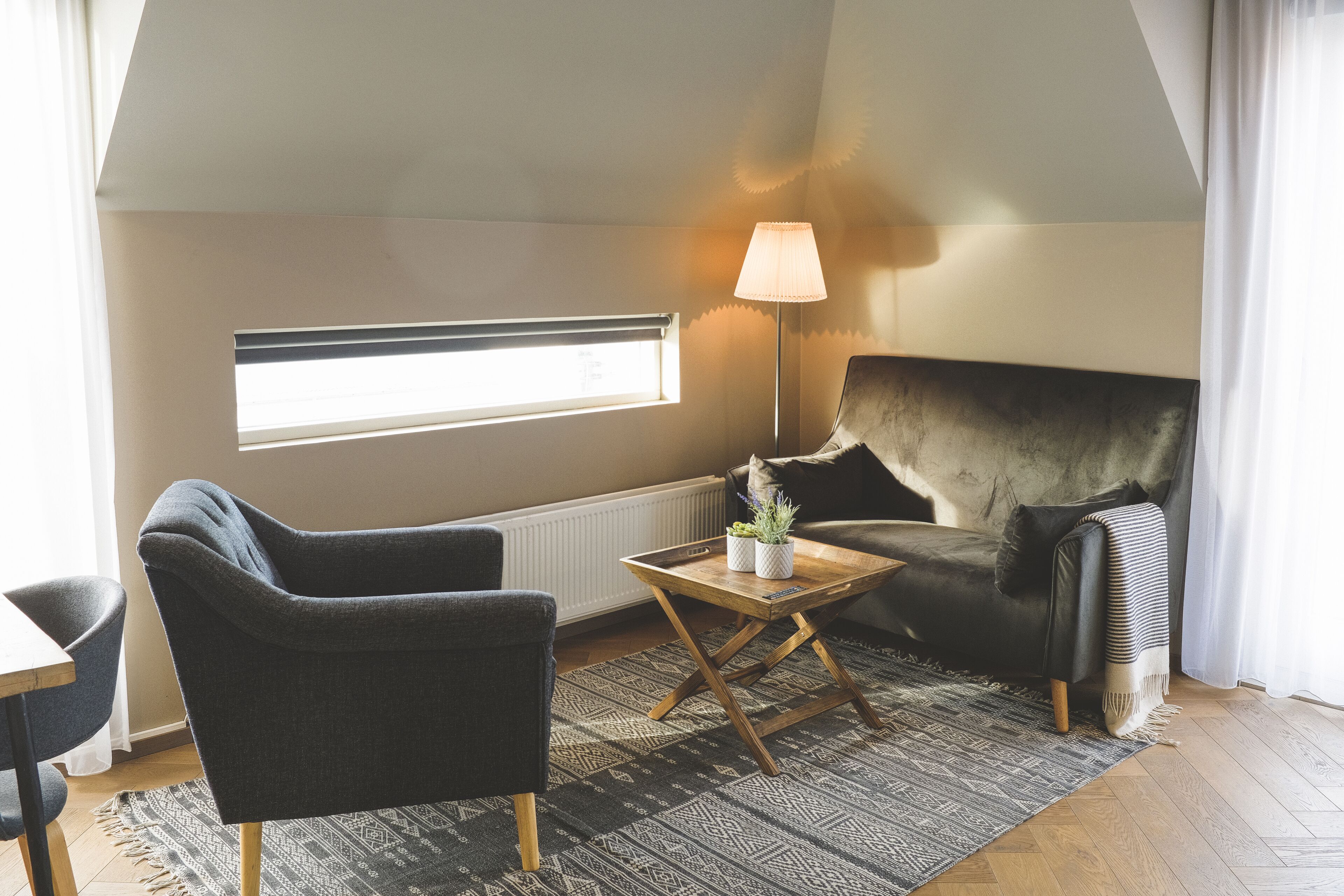 A cozy living room with a dark armchair, a sofa, a small wooden coffee table, and a floor lamp. There's a window with blinds and a radiator under the window.