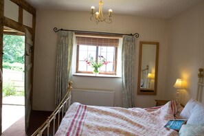 A bedroom with a bed, a window with curtains, a radiator, a chandelier, a bedside table with a lamp, and a mirror on the wall. - Tilbury Cottage (Taunton)