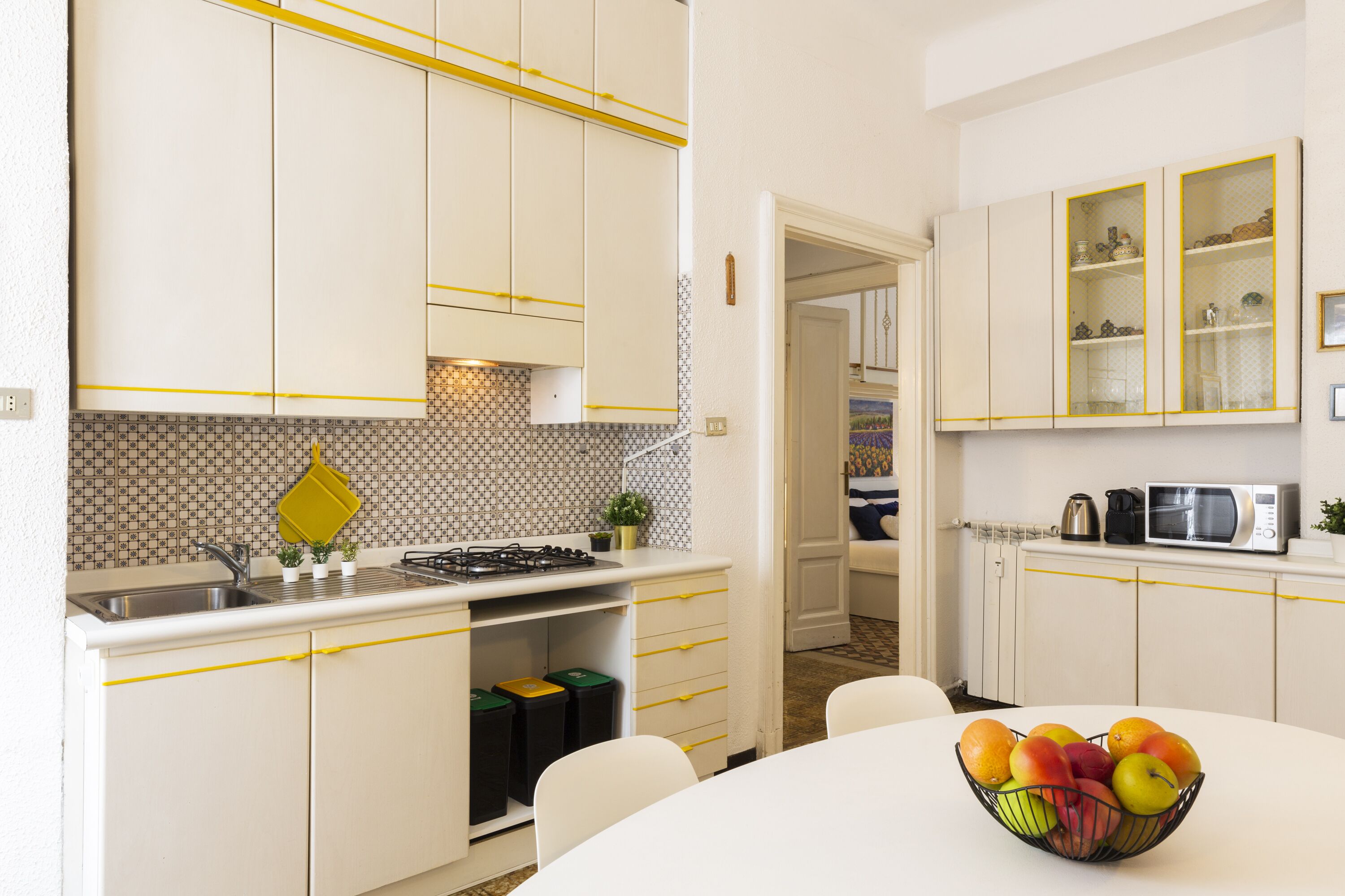 A kitchen with white cabinets, a sink, and a stove. There is a fruit bowl on the table.