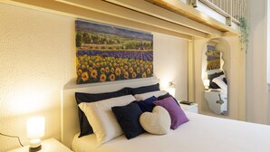 Ein Hotelzimmer mit Bett, Kissen, Nachttisch, Lampe und einem Gemälde einer Sonnenblumenwiese an der Wand.