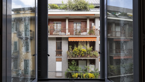 Blick durch ein Fenster auf ein Gebäude mit mehreren Balkonen, von denen einige mit Pflanzen bepflanzt sind, und einer sichtbaren Tür.