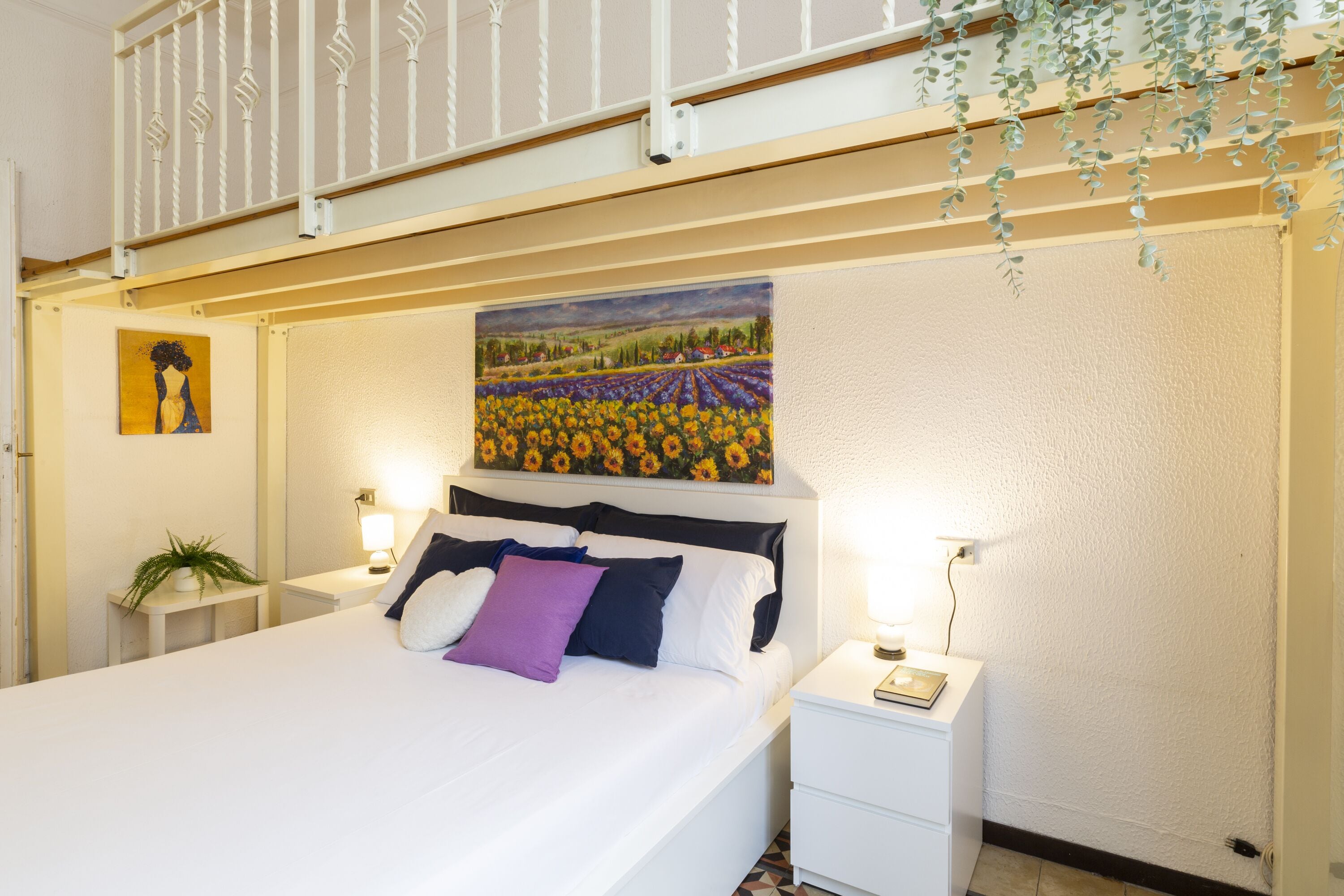 Un lit bien fait, avec un tableau représentant un champ de tournesols au-dessus, des tables de chevet blanches avec des lampes, et une petite plante sur le côté gauche.
