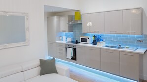 A modern kitchen with grey cabinets, a white countertop, and a built-in oven. There is a sink with a faucet, a microwave, and a kettle. The kitchen has a blue backsplash and a yellow exhaust hood. - Live in Leeds Greenhill Bungalows (Leeds)