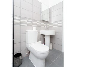 Bathroom features a white toilet, sink, and mirror, set against a backdrop of large, light-colored tiles.