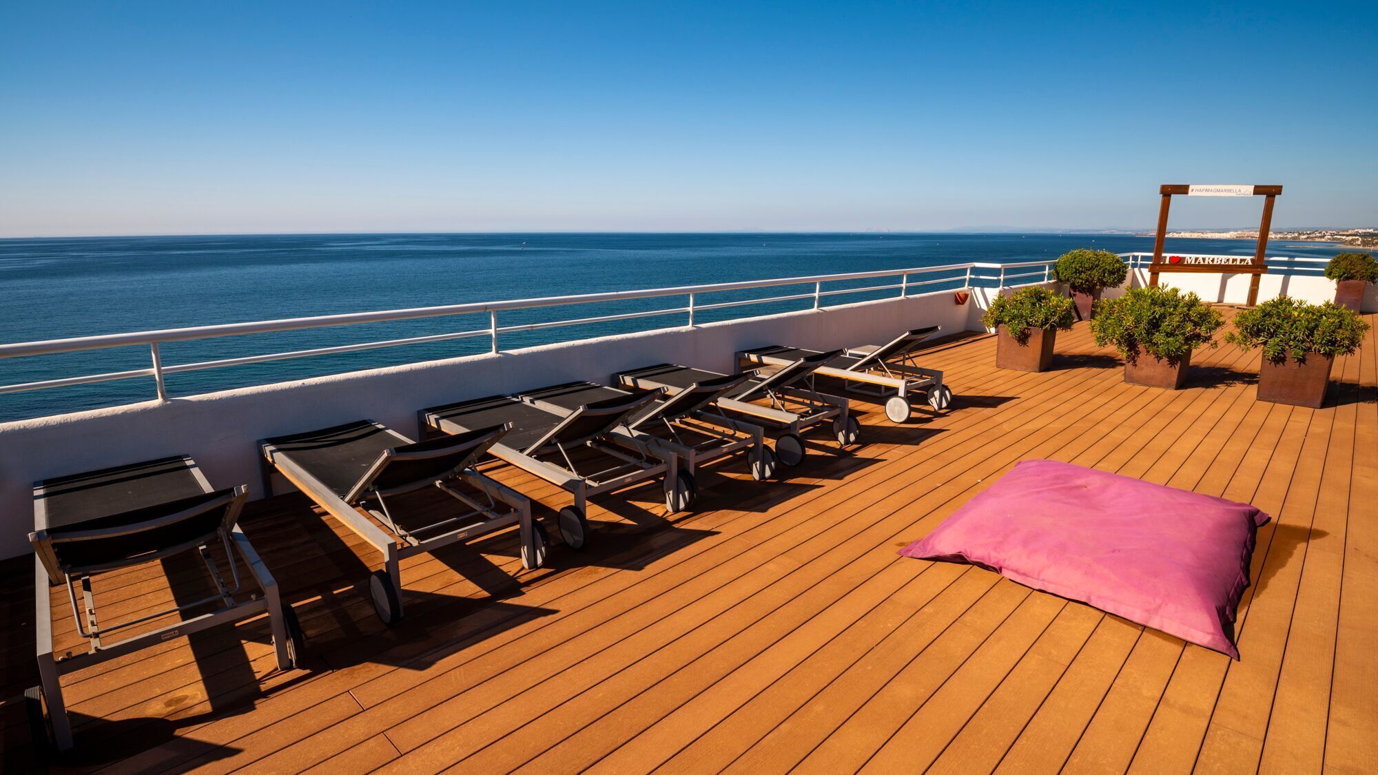 Terraza soleada con sillones de descanso y un cojín rosa, con vista al mar.