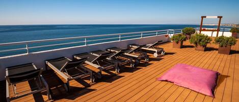 Terraza soleada con sillones de descanso y un cojín rosa, con vista al mar.