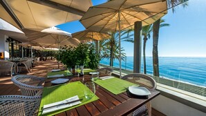 Un espace de restauration en plein air avec des tables en bois, des sets de table verts et des chaises en osier, offrant une vue sur l’océan.