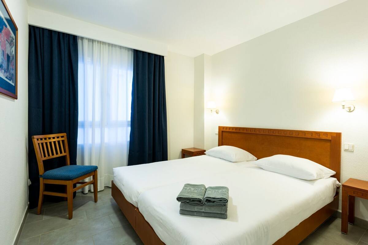 A hotel room with a bed, a chair, a nightstand, and a wall-mounted lamp.