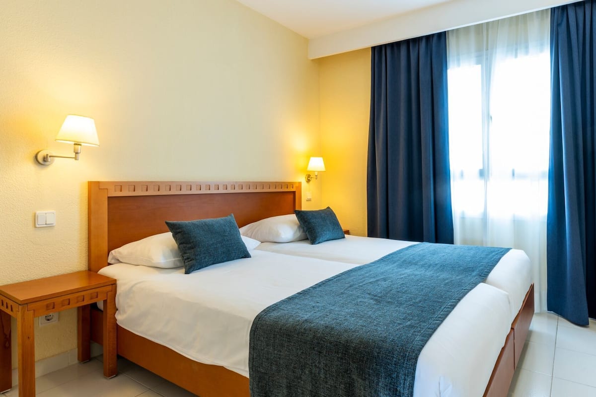 A double bed with a wooden headboard, two bedside lamps, a nightstand, and a large window with dark curtains.