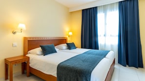 A double bed with a wooden headboard, two bedside lamps, a nightstand, and a large window with dark curtains. - Hapimag Resort Marbella (Marbella)