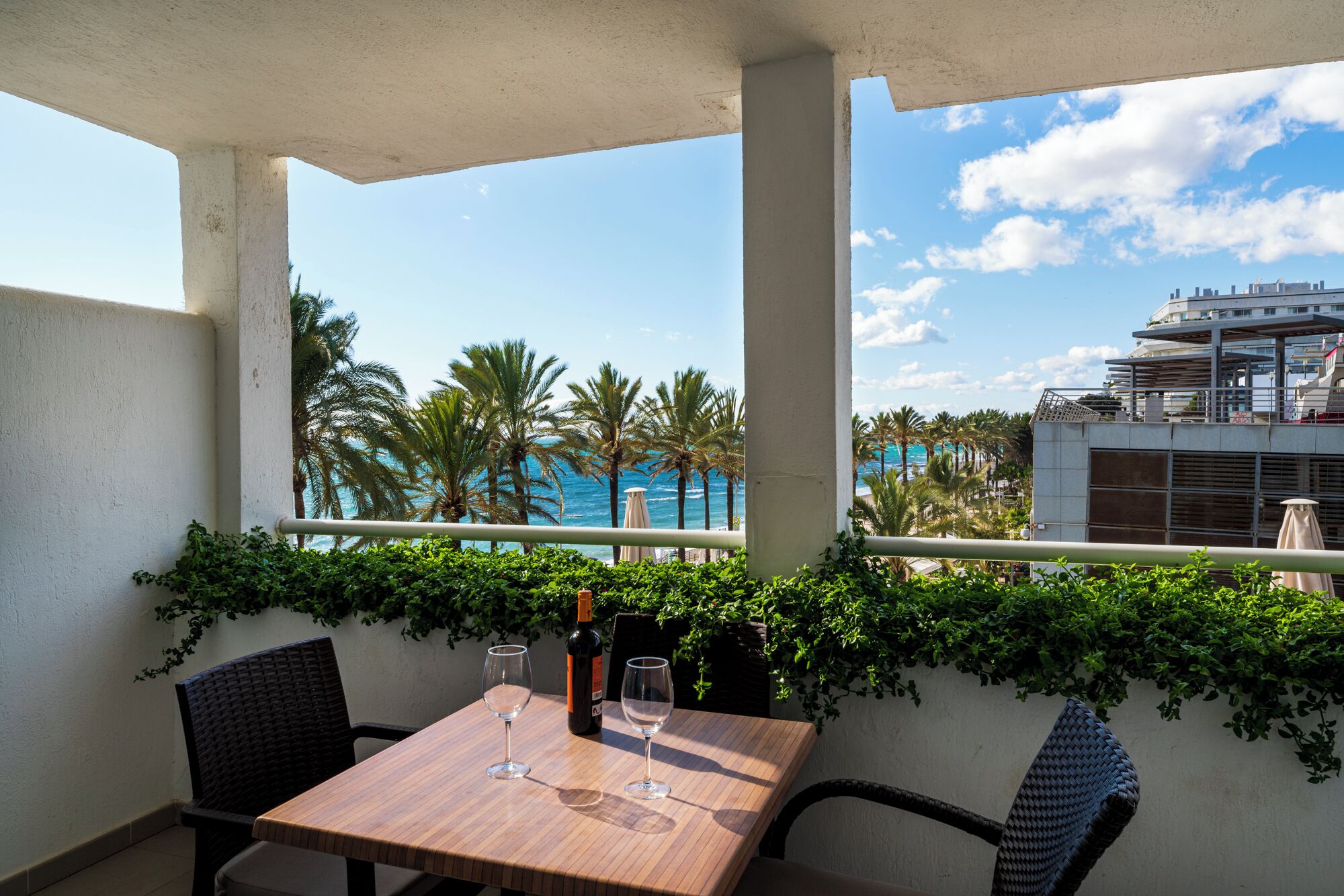 A balcony with a wooden table, two chairs, and a bottle of wine. There are palm trees and a view of the ocean and a building in the background.