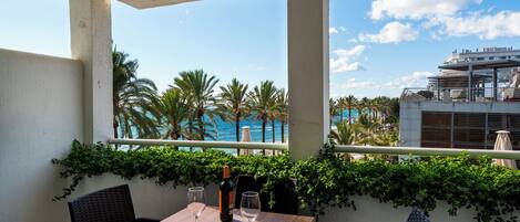 A balcony with a wooden table, two chairs, and a bottle of wine. There are palm trees and a view of the ocean and a building in the background.