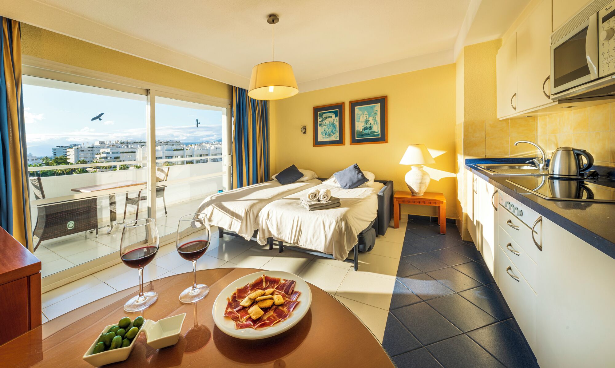 Une chambre d’hôtel avec un balcon, un lit, une table à manger avec des verres à vin et une assiette de nourriture, ainsi qu’un coin cuisine.