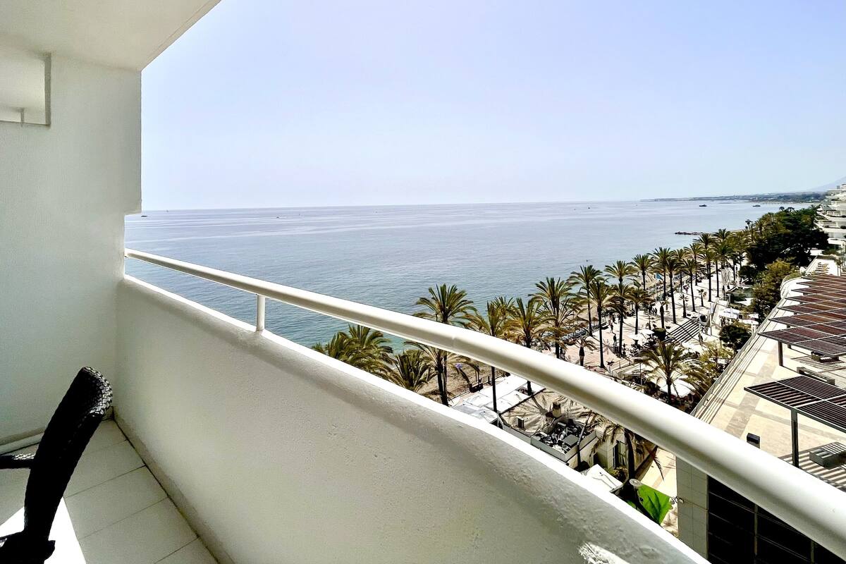 A balcony with a view of the sea and a beach area with palm trees and sun loungers.