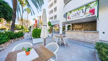 An outdoor cafe area with white wicker furniture, a wooden table, and a potted plant.
