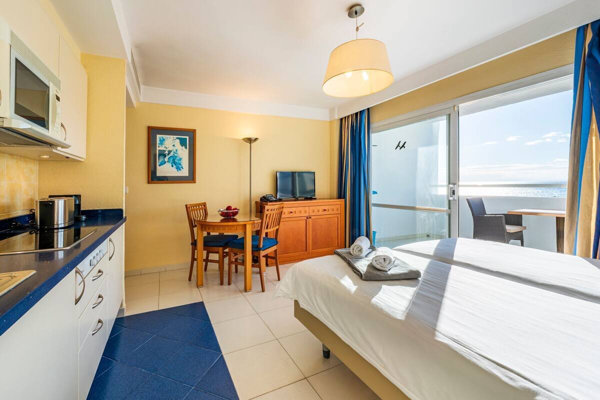 A hotel room with a bed, a desk with a chair, a kitchenette, and a view of the sea.
