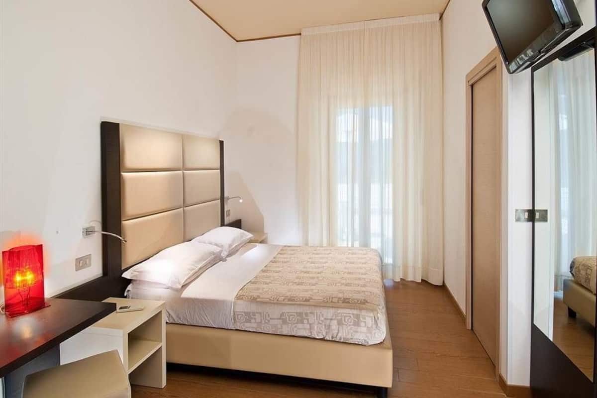 A hotel room with a bed, a desk with a lamp, a TV, and a window with curtains.