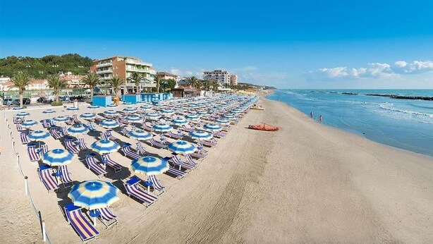 Ein Strand mit Reihen von Sonnenschirmen und Liegestühlen, einem klaren Himmel und Gebäuden im Hintergrund.