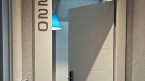 Ein Flur mit einer verschlossenen Tür mit der Aufschrift 'ONNO', die zu einem Zimmer mit einem Bett und einer blauen Lampe führt.