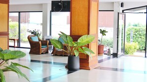 A spacious lobby with a checkered floor, a large potted plant, a wicker chair, and a view of the outdoors through large windows. - Bordin Hotel (Ubon Ratchathani)