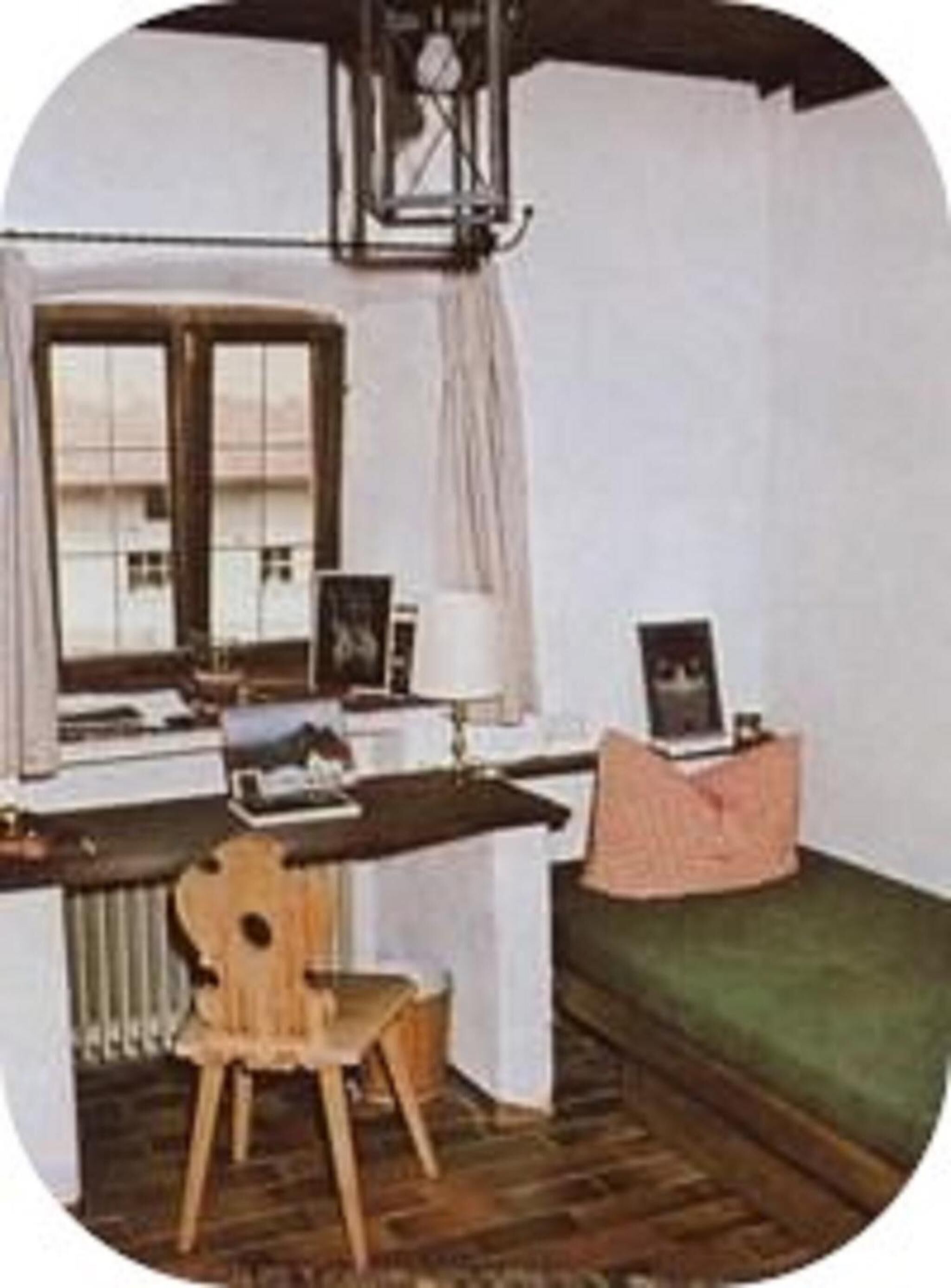 Una habitación con escritorio de madera, una silla, cama con cubierta verde, ventana con cortinas y un radiador.
