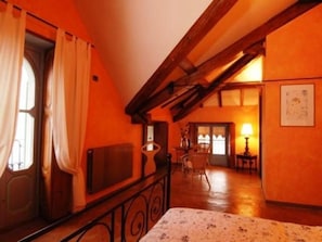 A room with a sloped ceiling, a bed with a floral bedspread, a radiator, a chair, and a table with a lamp.