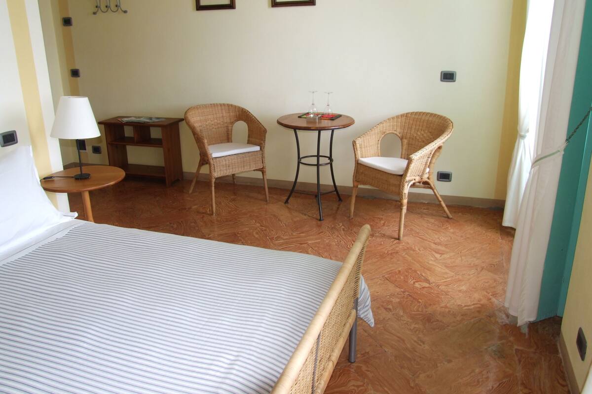 A hotel room with a bed, two wicker chairs, a small table, a lamp, and two framed pictures on the wall.